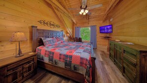 Camera da letto in una baita di legno con un letto, una televisione, una ventola a soffitto e una finestra con tende.