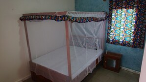 A single bed with a mosquito net, a small wooden stool, and a colorful stained glass window. - LA CASE A MAMY (Nianing)