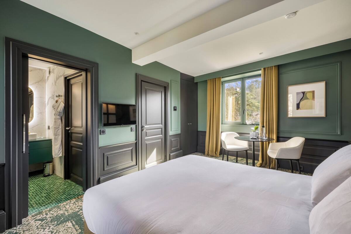 A hotel room with a bed, a desk, two chairs, a TV, and a window with curtains.