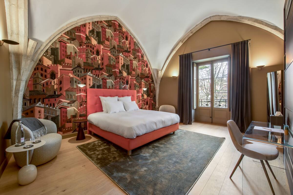 A bedroom with a red bed, a glass table, and a cityscape mural on the wall.