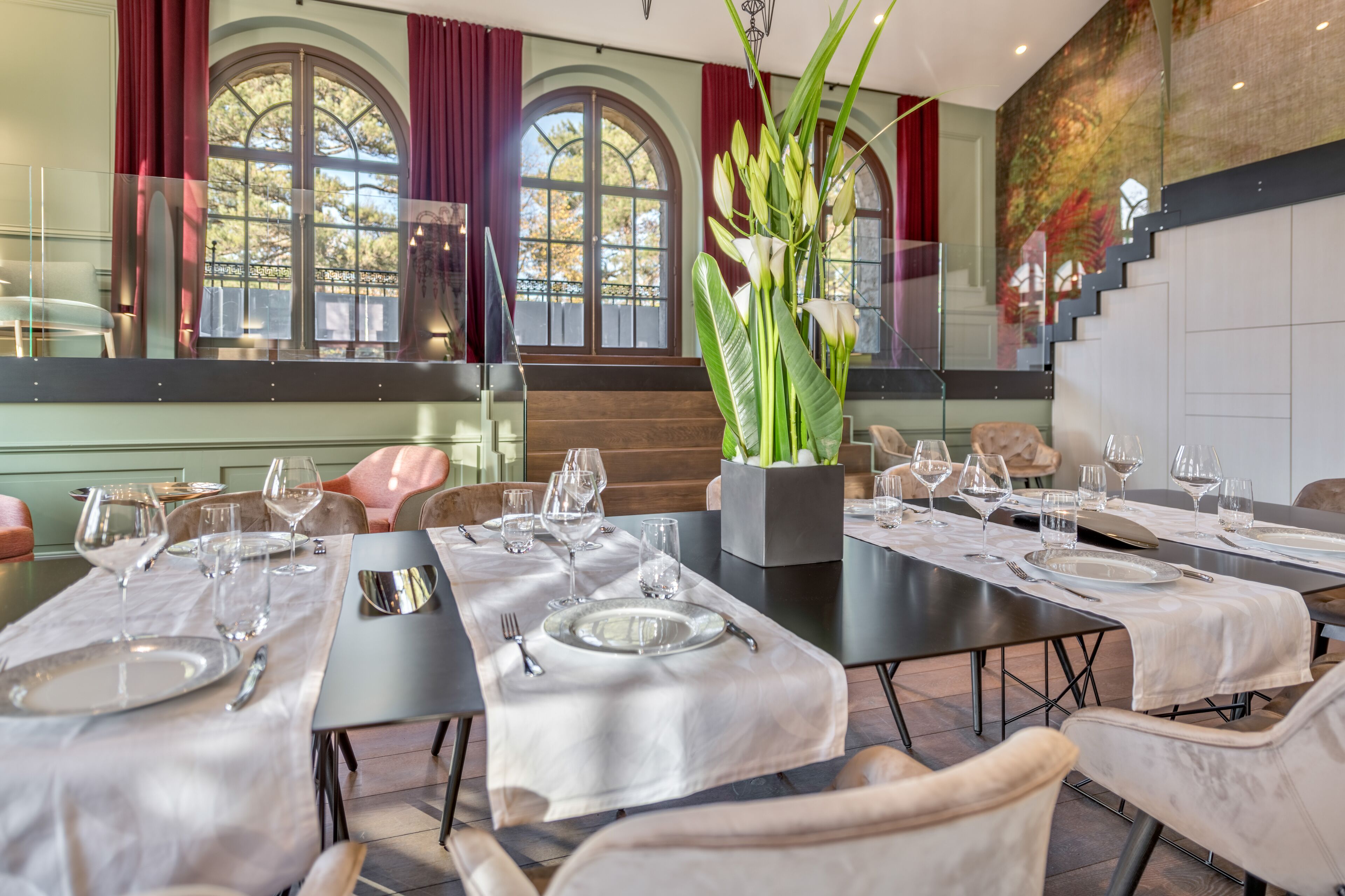 Un comedor con mesas dispuestas para una comida, ventanas grandes con cortinas y una escalera al fondo.