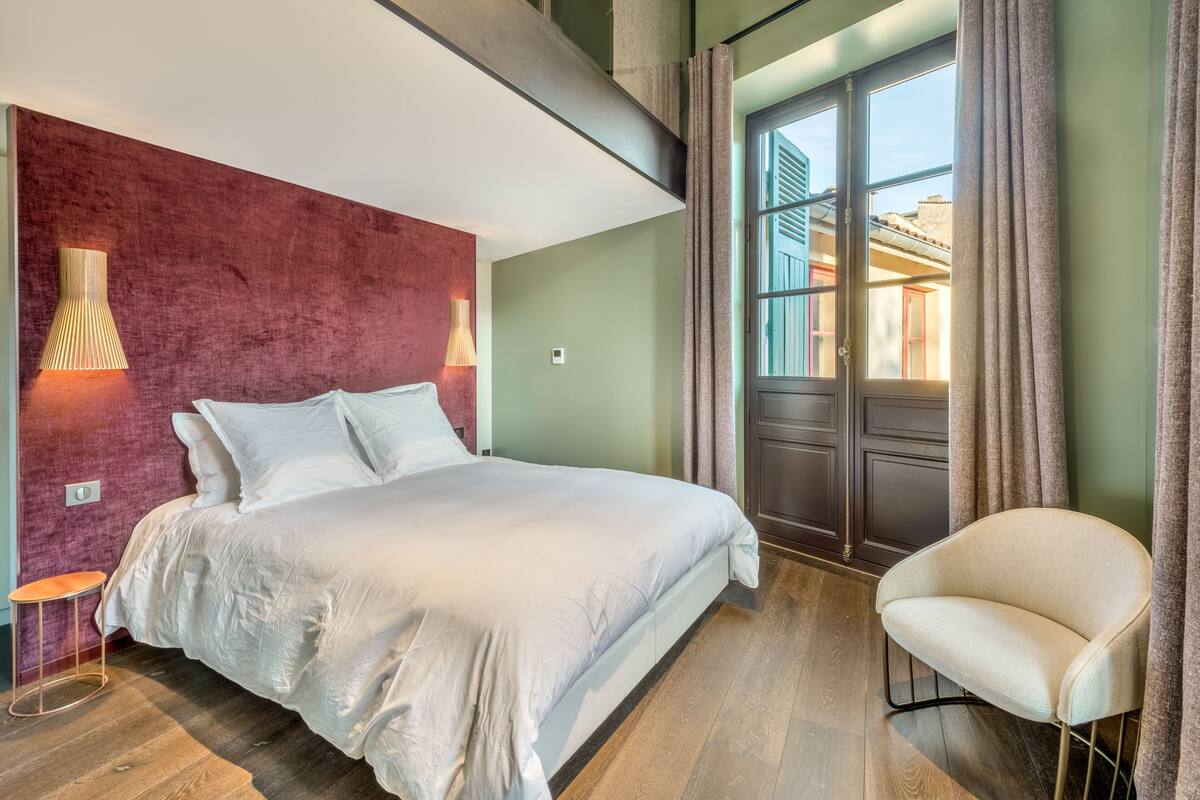 A modern bedroom with a large bed, a chair, and a view of the outside through a window.