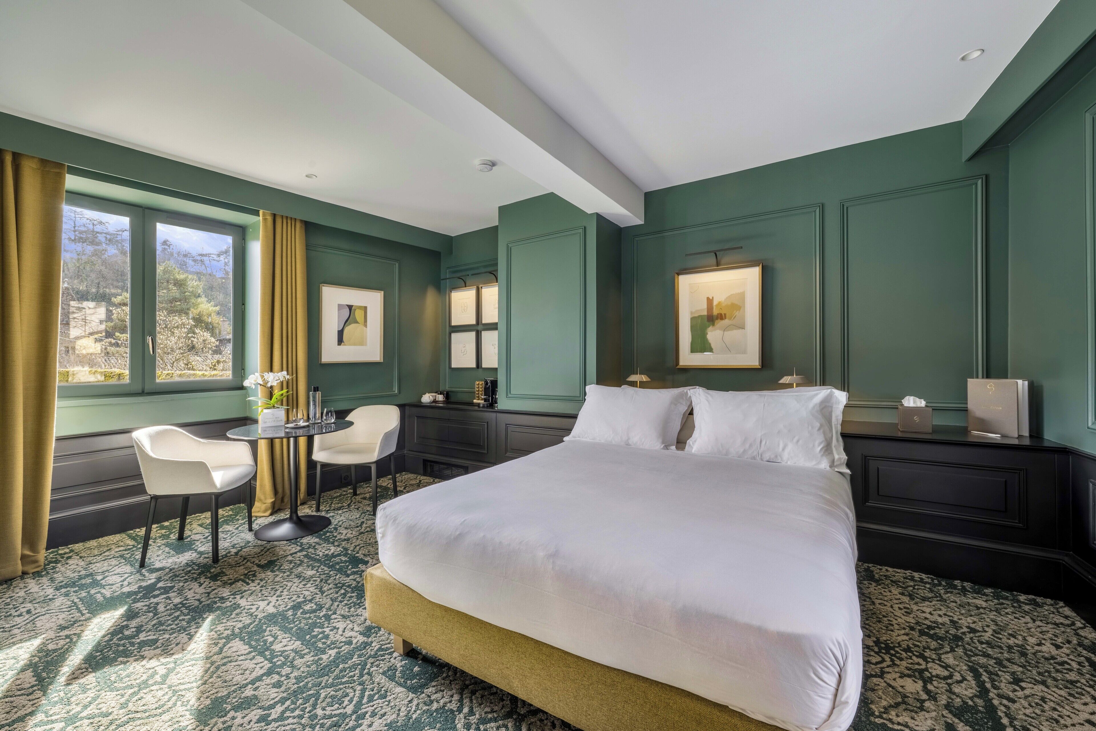 Habitación de hotel con una cama grande, un escritorio con silla, una ventana con cortinas y obras de arte enmarcadas en las paredes.