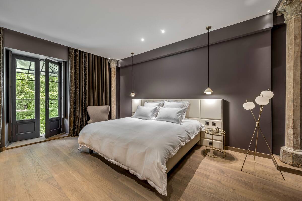 A modern bedroom with a large bed, a nightstand, a lamp, and a view of greenery outside.