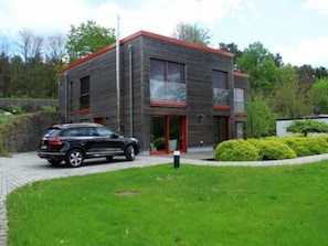 A modern two-story building with a brick facade and red accents, a paved driveway, and a well-maintained lawn. - Vacation apartment IV Sunset (Mittenwalde)
