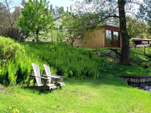 A grassy area with two Adirondack chairs, a small wooden structure, and a pond. - Vacation apartment IV Sunset (Mittenwalde)