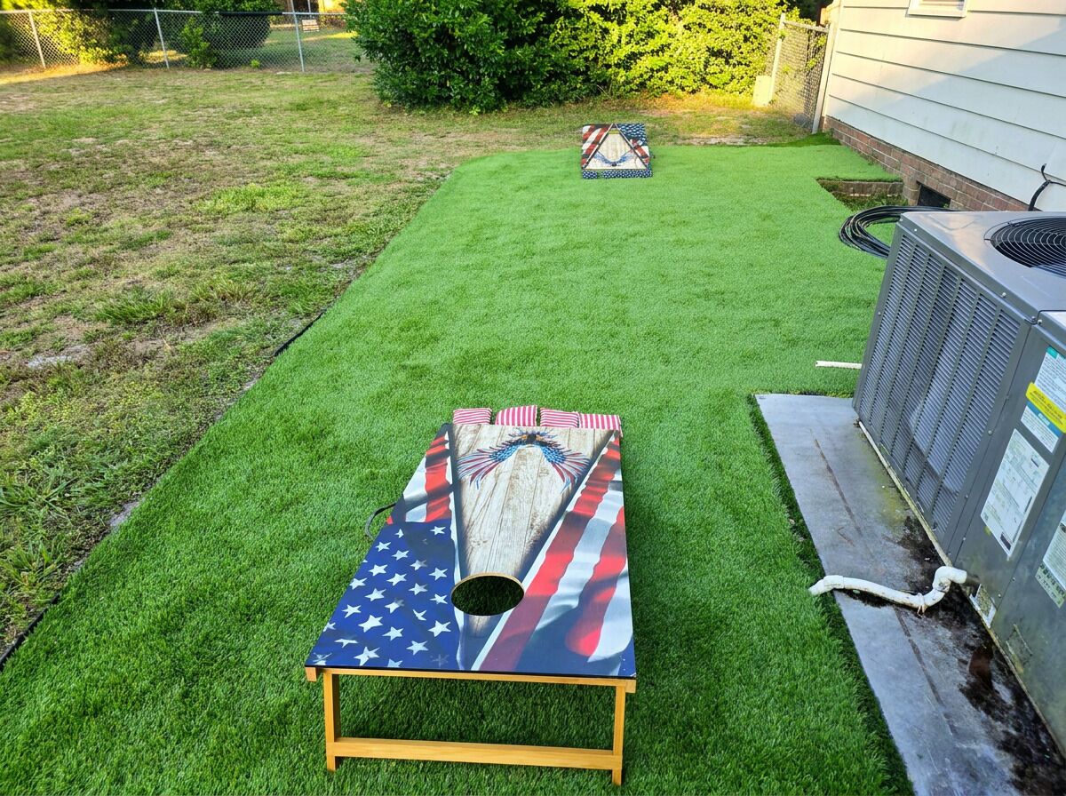 Un patio con césped artificial, una mesa de madera con diseño de bandera estadounidense y una unidad de aire acondicionado.