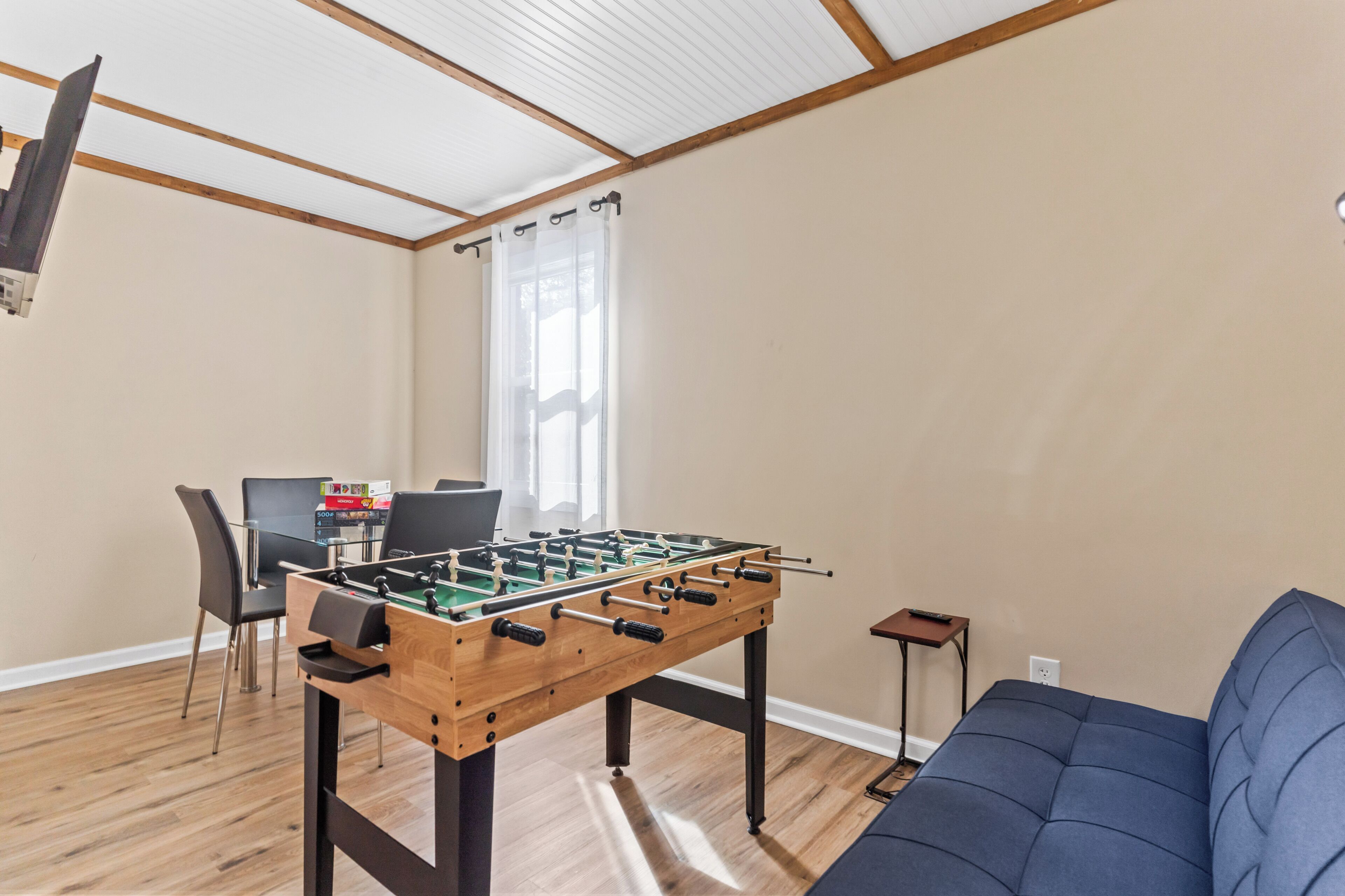 A room with a foosball table, a sofa, a dining table with chairs, and a window with curtains.