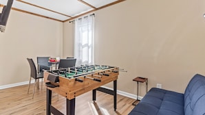Une pièce comprenant une table de baby-foot, un canapé, une table à manger avec des chaises et une fenêtre avec des rideaux.