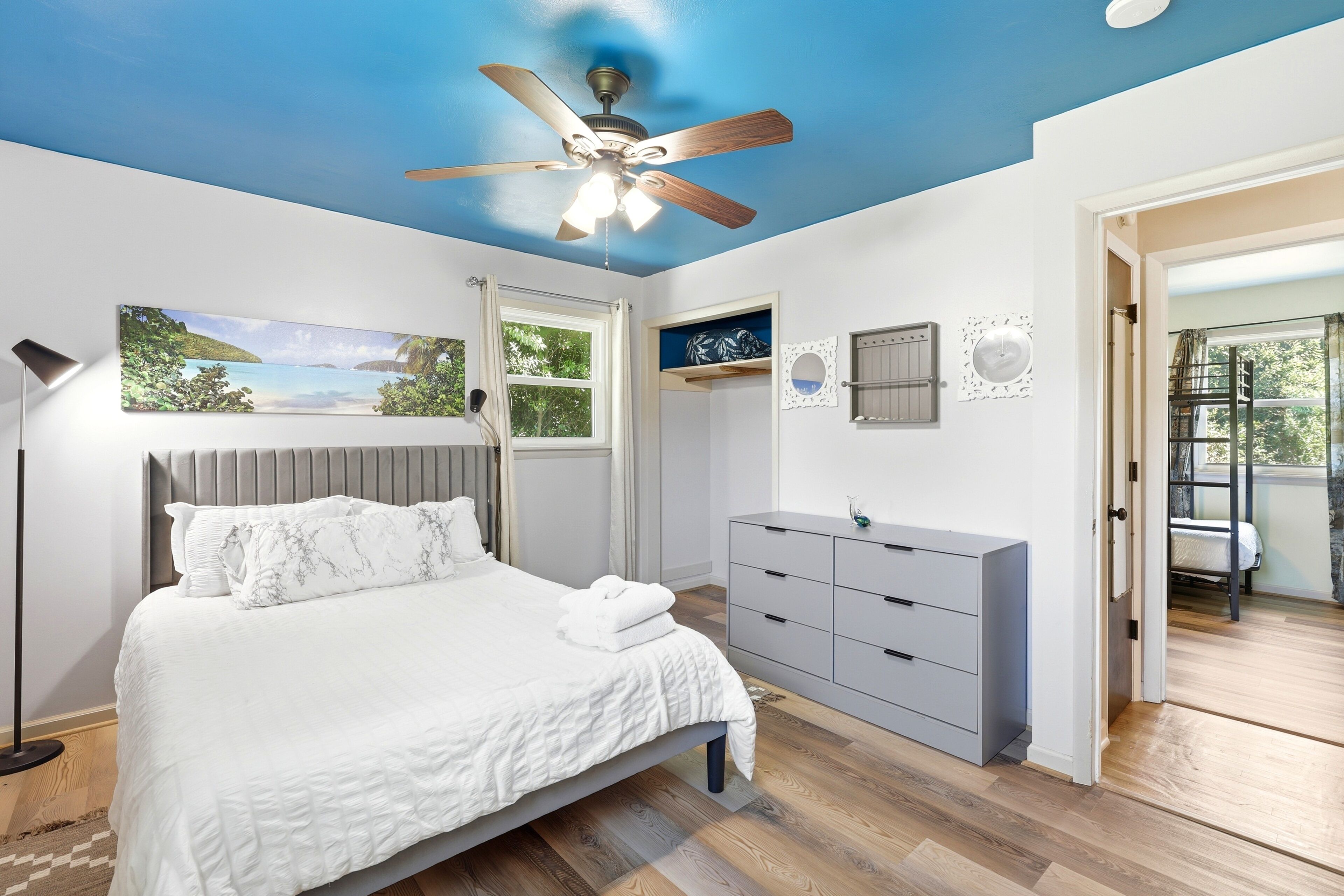 Ein Schlafzimmer mit Bett, Kommode, Deckenventilator, Fenster und einer Tür, die auf einen Balkon führt.