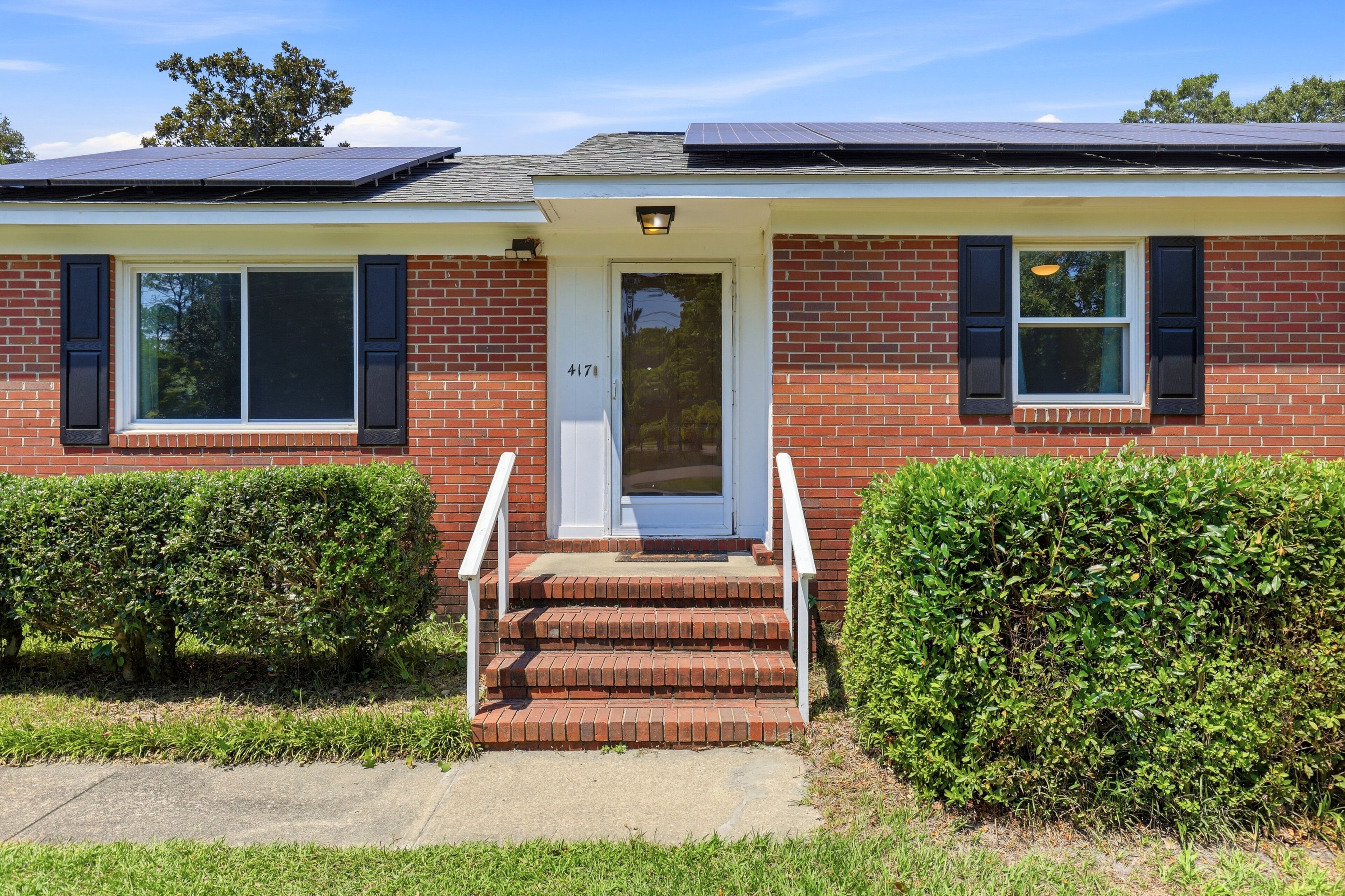 A two-story house with a red brick exterior, a white front door, and solar panels on the roof.