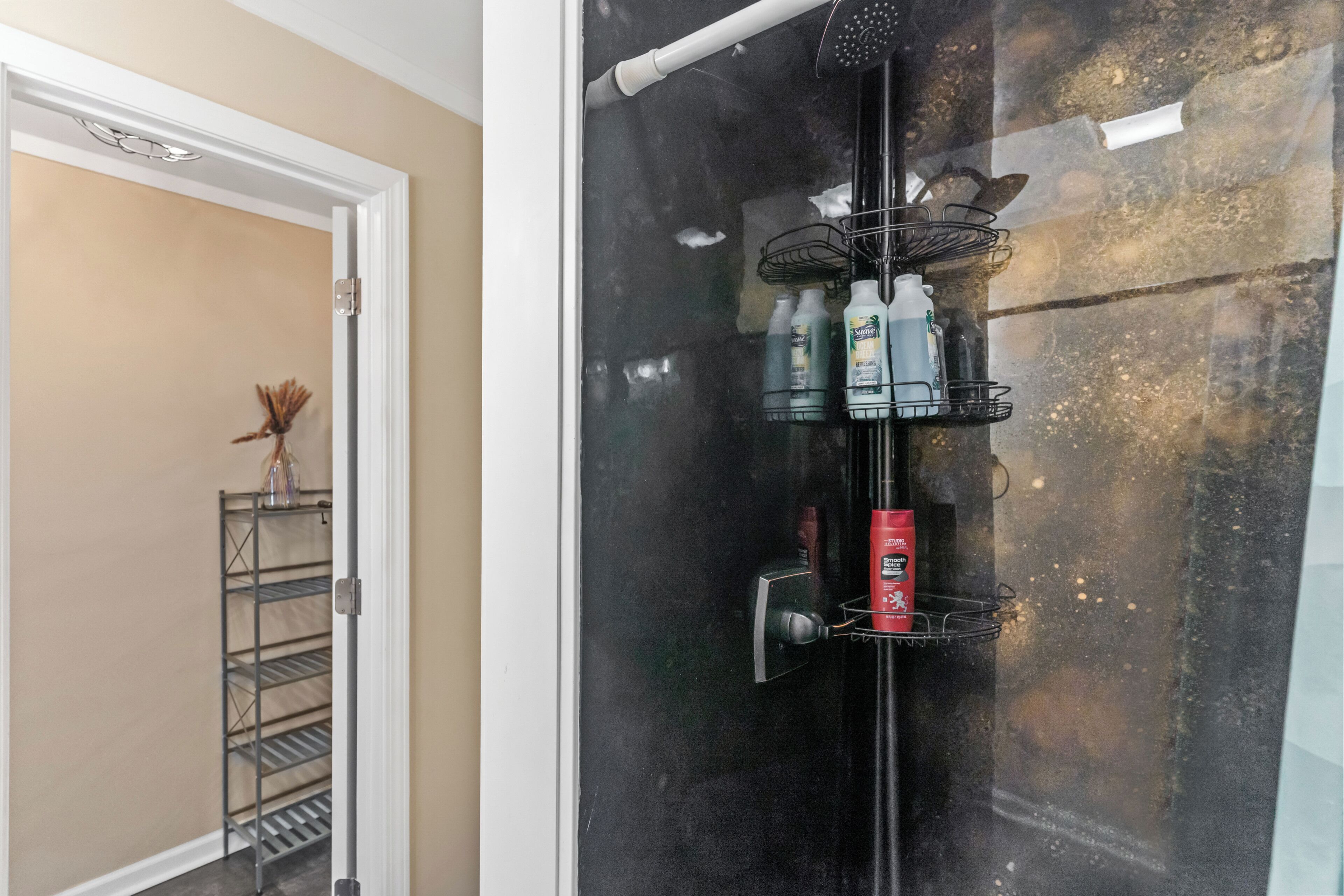 A bathroom with a glass shower enclosure, a metal shelf with toiletries, and a door leading to another room.