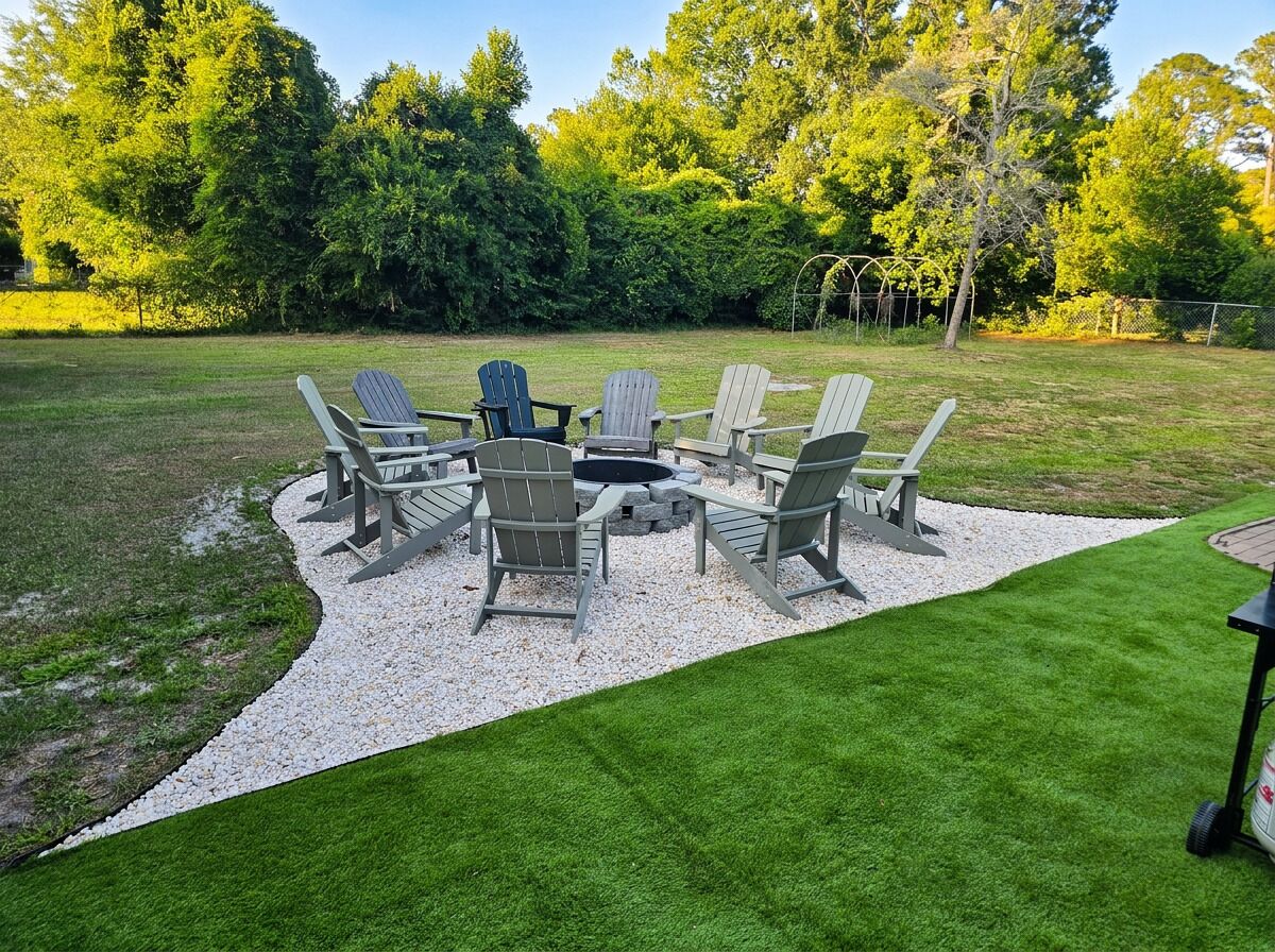 En terrasse med Adirondack-stole og et bord, omgivet af en velplejet græsplæne og træer.