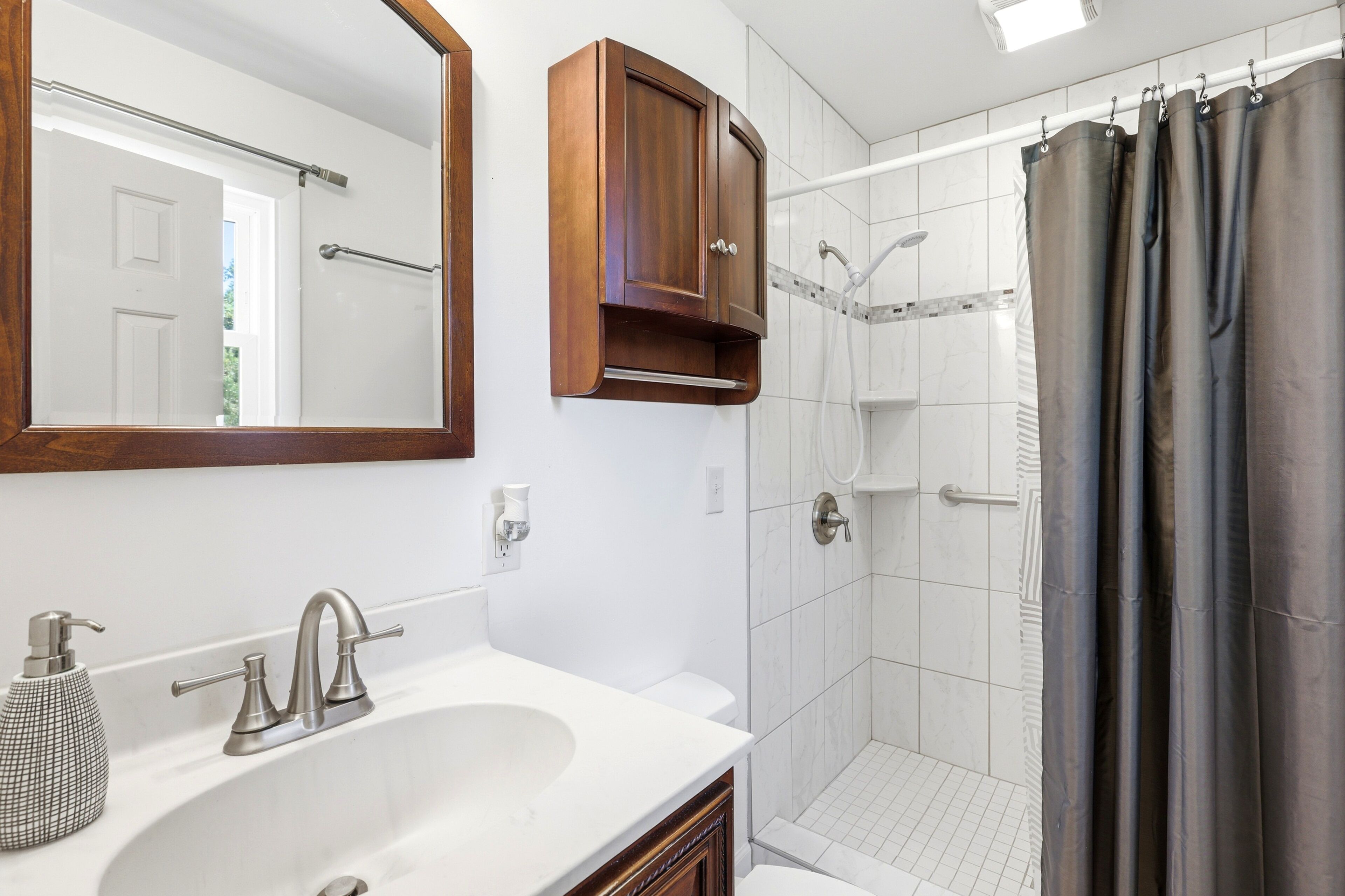 Un baño con lavamanos blanco, espejo, mueble de madera y zona de ducha con cortina.