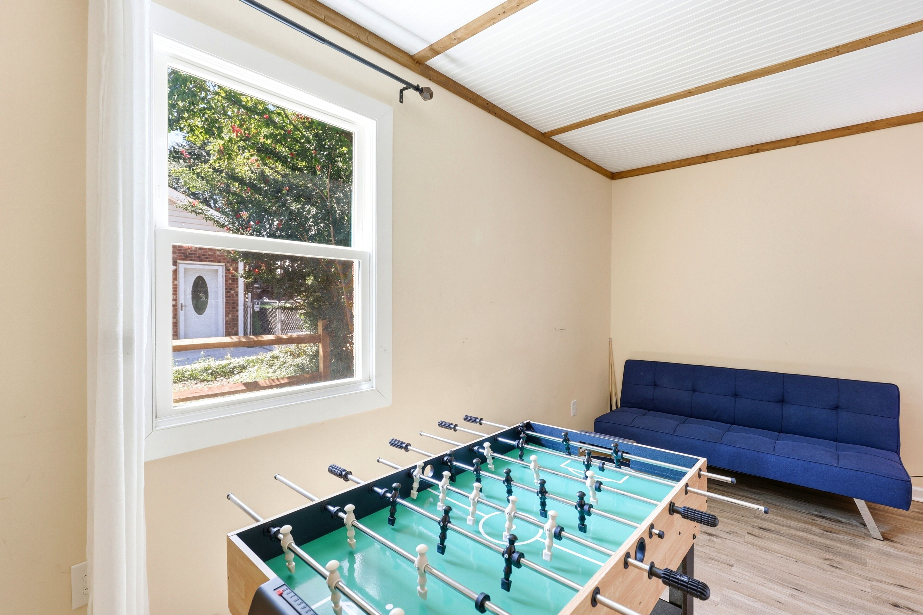 Ein Zimmer mit einem Kicker, einem Fenster mit Blick auf einen Baum und ein Gebäude, sowie ein blaues Sofa.