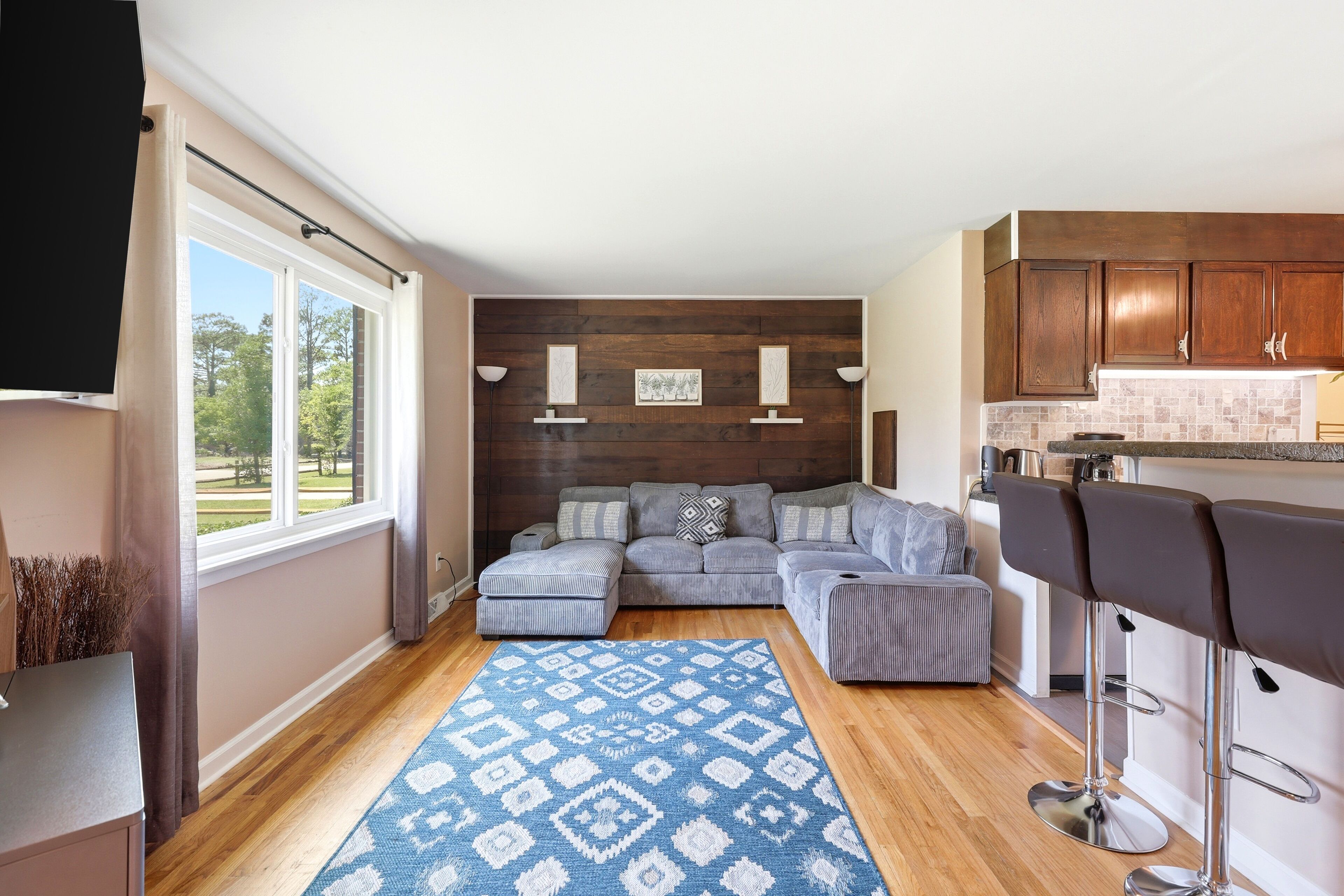 A modern living room with a sectional sofa, a patterned rug, and a kitchen area with wooden cabinets and a bar stool.