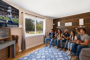 A living room with a large window, a television displaying a football game, and a group of people sitting on a couch.