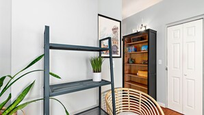 Ein Zimmer mit einem Metallregal, einem Korbstuhl, einem Bücherregal und einem gerahmten Bild an der Wand.