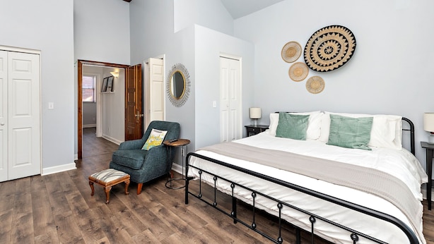 Ein Schlafzimmer mit einem großen Bett, einem Sessel, einem kleinen Hocker und einem Spiegel an der Wand.