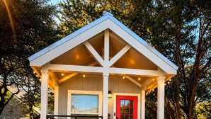 A small wooden structure with a red door, a porch with outdoor seating, and a gravel ground. - Poppy Seed Cottage (Canyon Lake)