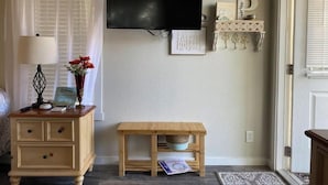 A room with a wooden nightstand, a small table, a TV mounted on the wall, a decorative shelf, and a door with a window. - Poppy Seed Cottage (Canyon Lake)