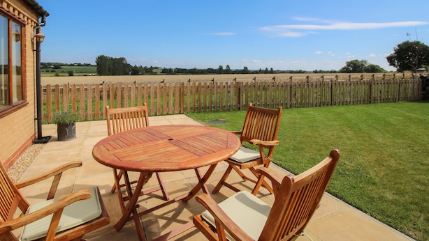 Une terrasse avec une table ronde et six chaises en bois, une clôture en bois et, au fond, un champ d’herbe.