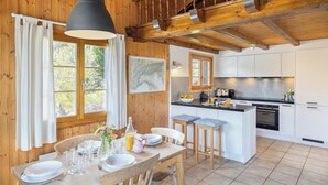 A wooden cabin with a dining area, kitchen, and a large window with curtains. - Chalet Les Crus (Riddes)
