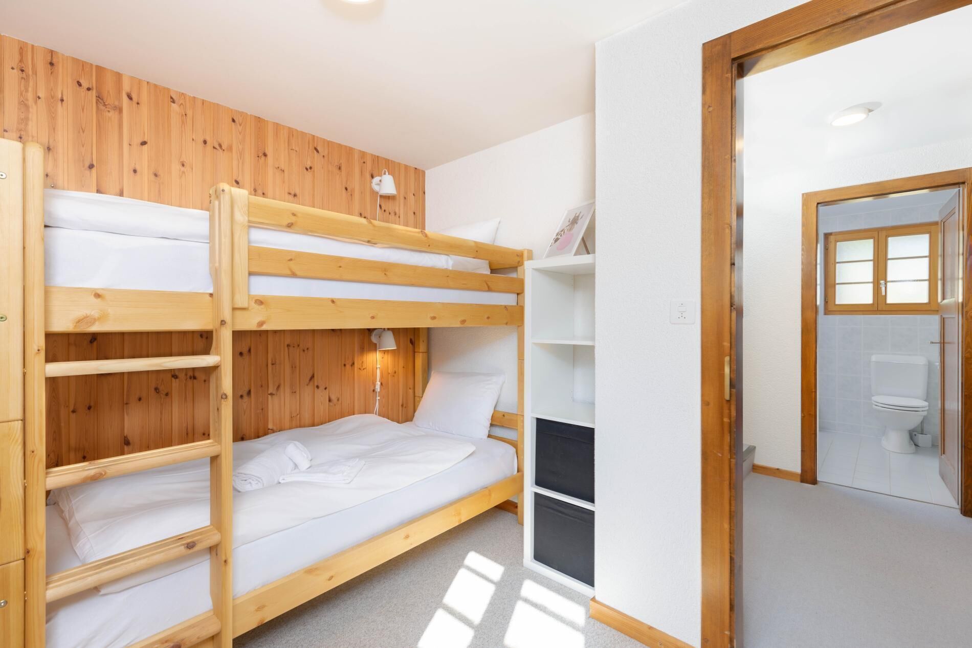 Une chambre avec des lits superposés, des murs en bois, une porte blanche et une salle de bain équipée de toilettes et d’un lavabo.