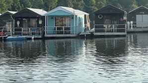 A row of floating houses with a calm water surface in the foreground and a forested hill in the background.