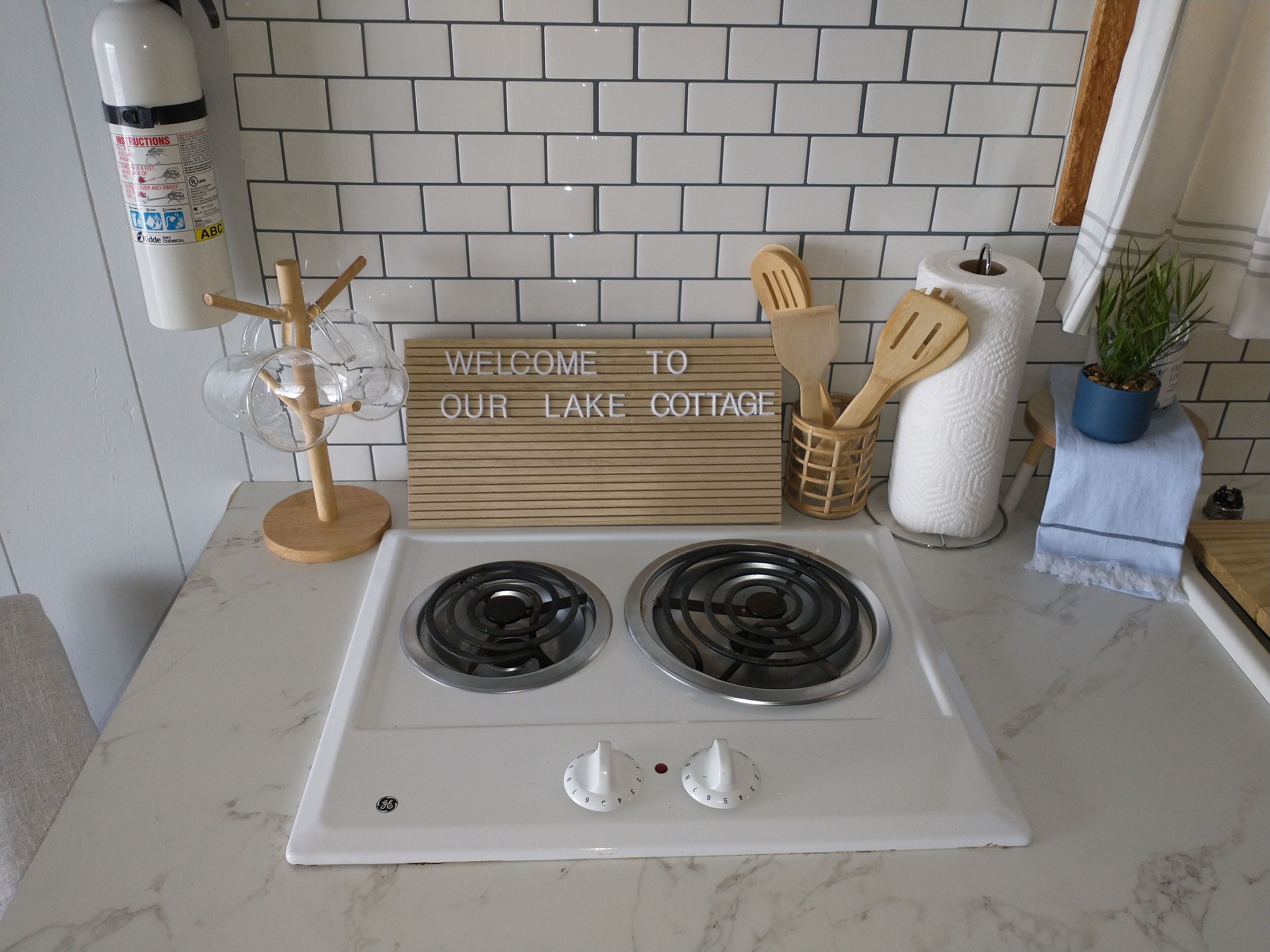 Uma cozinha com fogão branco, uma placa com a inscrição 'WELCOME TO OUR LAKE COTTAGE', utensílios de madeira e um vaso com planta.