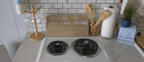 Uma cozinha com fogão branco, uma placa com a inscrição 'WELCOME TO OUR LAKE COTTAGE', utensílios de madeira e um vaso com planta.