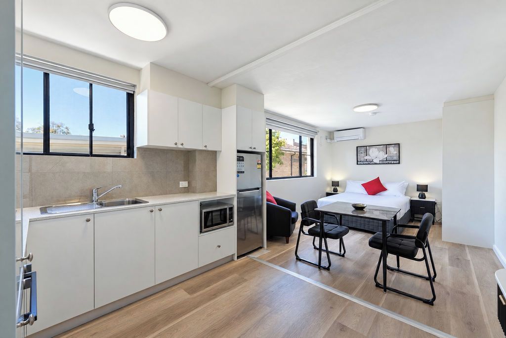 Een moderne keuken met een inbouwmagnetron, een koelkast en een spoelbak. Er is een zithoek met een tafel en stoelen, en een bed met een rood kussen.