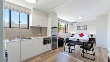 Een moderne keuken met een inbouwmagnetron, een koelkast en een spoelbak. Er is een zithoek met een tafel en stoelen, en een bed met een rood kussen.