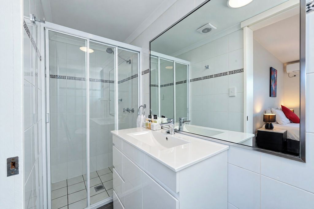 A modern bathroom with a glass-enclosed shower, a white sink, and a large mirror reflecting a bed with a red pillow.