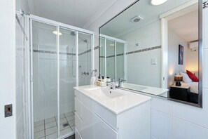 Ein modernes Badezimmer mit einer Glaskabine zur Dusche, einem weißen Waschbecken und einem großen Spiegel, der ein Bett mit einem roten Kissen reflektiert.