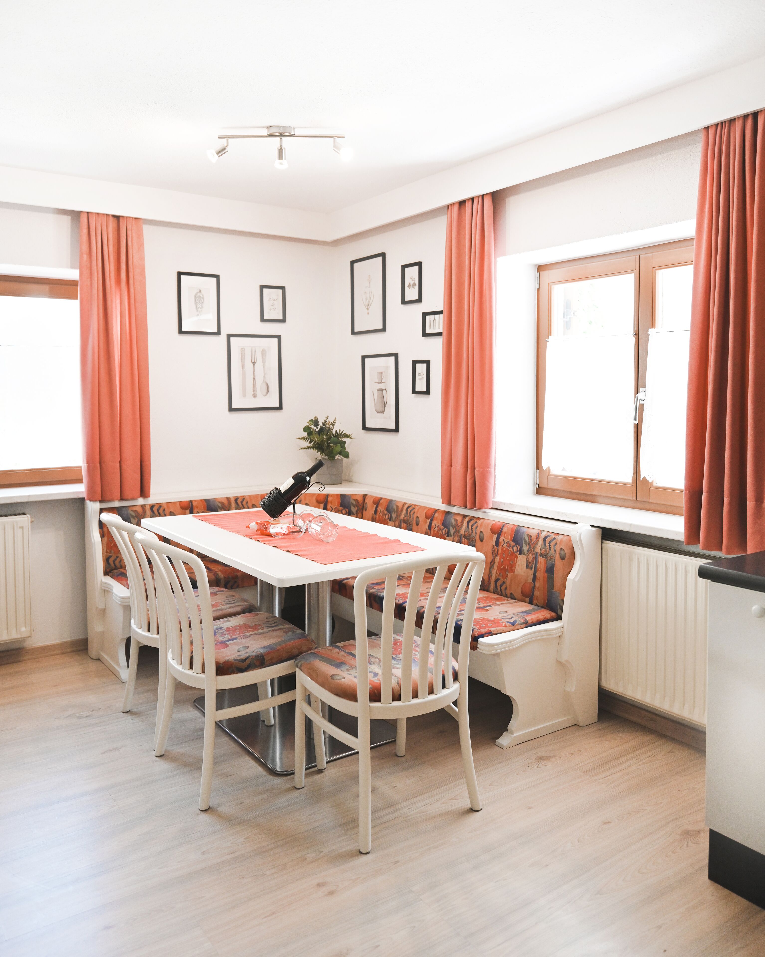 Un coin repas avec une table et des chaises blanches, un banc agrémenté de coussins à motifs, des tableaux encadrés au mur et une fenêtre avec des rideaux.