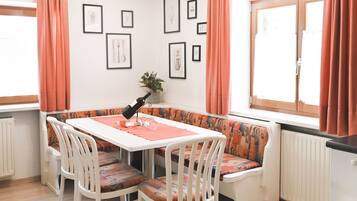 Un coin repas avec une table et des chaises blanches, un banc agrémenté de coussins à motifs, des tableaux encadrés au mur et une fenêtre avec des rideaux.