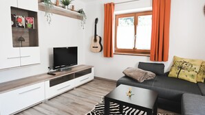 Ein modernes Wohnzimmer mit einem Flachbildfernseher, einer Gitarre, einer Couch mit Kissen, einem Teppich mit Zebra-Muster und einem Fenster mit orangefarbenen Vorhängen.
