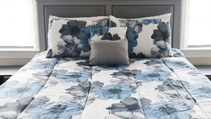 Ein Bett mit einem blumigen Bettbezug und zwei Kissen, eines mit floralem Muster und eines einfarbig, in einem Zimmer mit dunklem Kopfteil und einem Fenster mit Jalousien.