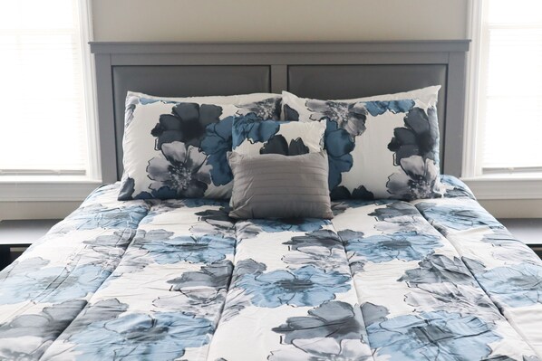 Una cama con una colcha estampada con motivos florales y dos almohadas, una con diseño floral y otra de color liso, en una habitación con cabecera oscura y una ventana con persianas.
