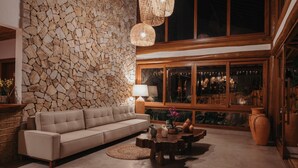 Ein gemütliches Wohnzimmer mit Steinwand, einem beigefarbenen Sofa, einem Holztisch und einem großen Fenster mit Blick auf den Nachthimmel.
