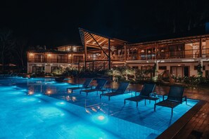 Area piscina di notte, con poltrone relax e un edificio con numerosi balconi.
