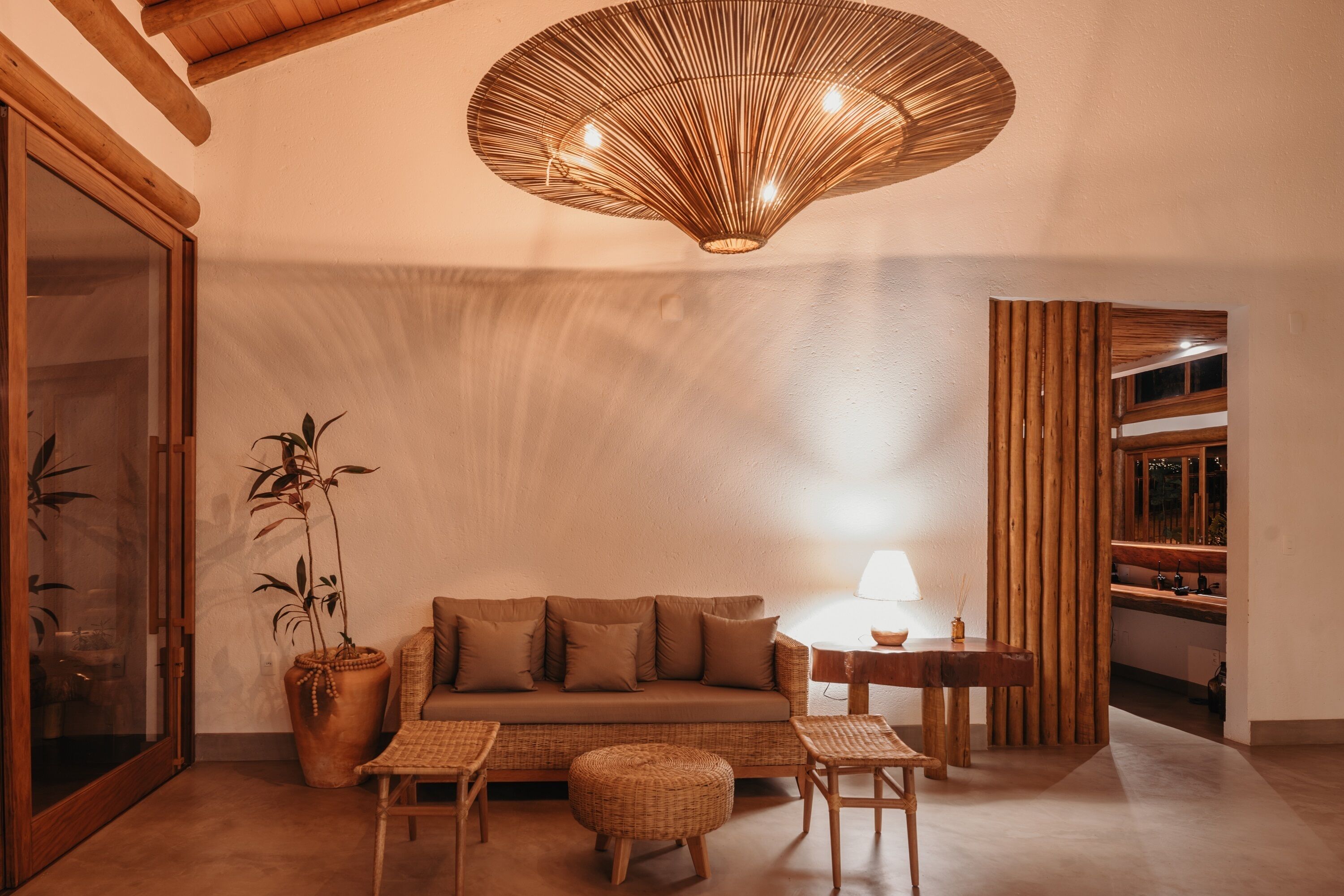 A cozy living room with a sofa, ottoman, and wooden side tables. A large woven light fixture hangs from the ceiling, and a potted plant is on the floor.