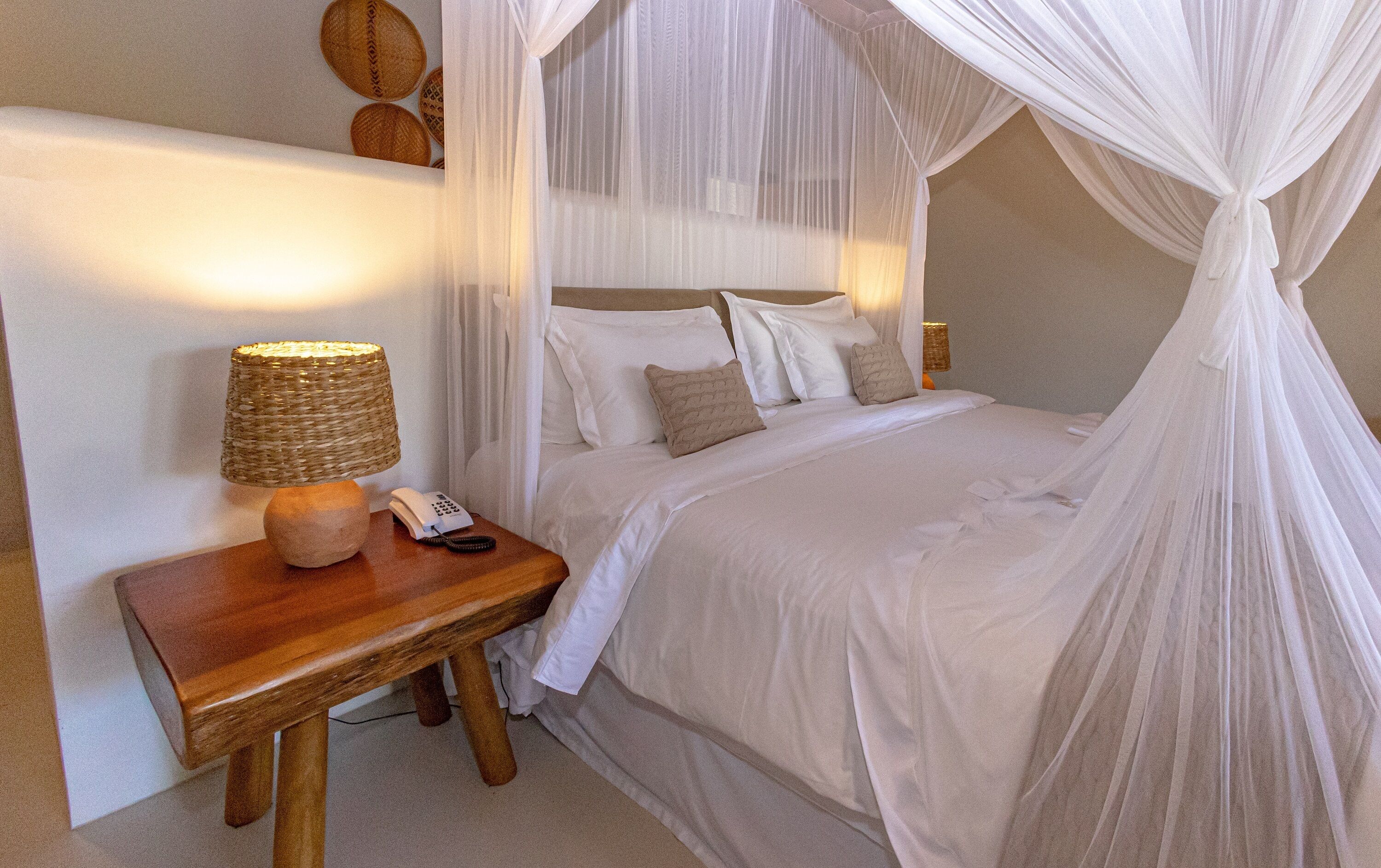 A neatly made bed with white linens and a canopy, a bedside lamp, a wooden nightstand, and a telephone.