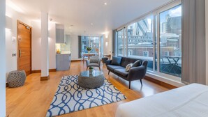 A modern hotel room with a large window, a sofa, a dining area, and a kitchen in the background. - Tower Hill City Centre Luxury Apartments (London)