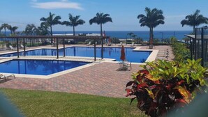 Zona de piscina con vista al océano, rodeada de palmeras y sillones de descanso.