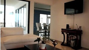 Una sala moderna con un sofá, una mesa de centro con una planta, un televisor montado en la pared y un comedor al fondo.