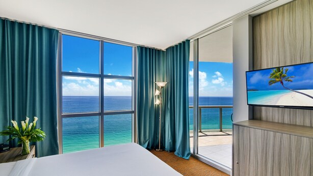 Une chambre d’hôtel dotée d’une grande fenêtre donnant sur l’océan, d’un lit, d’un téléviseur posé sur une commode en bois et d’un lampadaire.