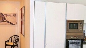 Eine kompakte Küche mit integriertem Backofen und Mikrowelle in einem weißen Schrank, ein Stuhl und ein gerahmtes Kunstwerk an der Wand.