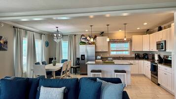 A modern living room with a dark blue sofa, a dining area with a white table, and a kitchen with white cabinets and stainless steel appliances.
